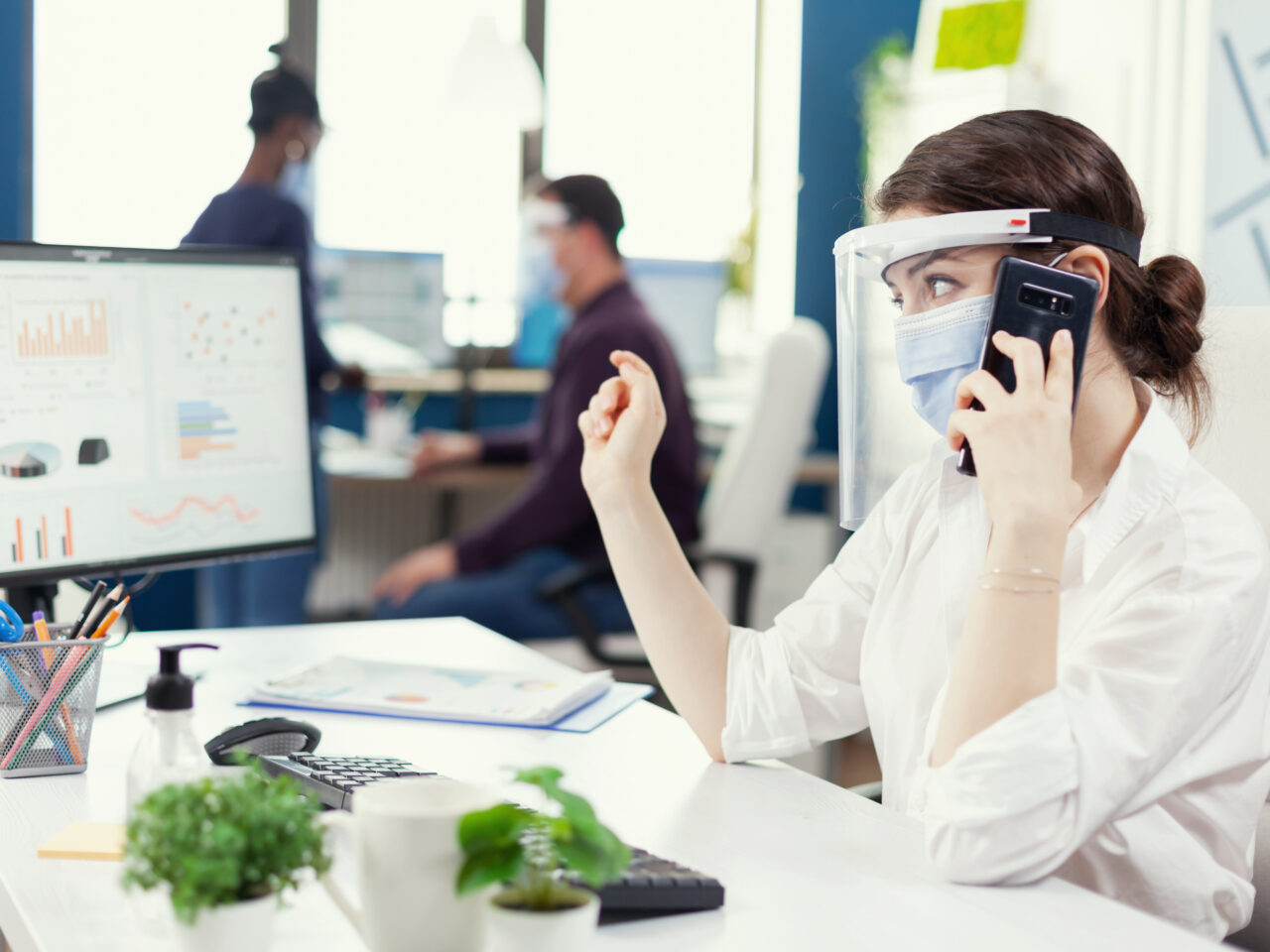 Business administrator sitting at her workplace wearing face mask during covid19 talking on smartphone. Multiethnic coworkers working respecting social distance in financial company.