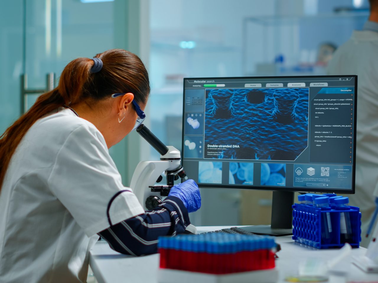 Biotechnology scientist researching in laboratory using microscope and typing on pc. Chemist examining virus evolution using high tech for scientific research of vaccine development against covid19