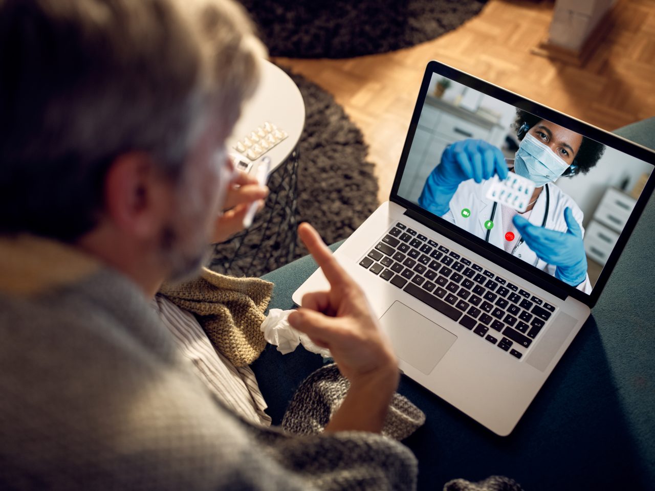 Close-up of man with fever consulting his doctor during a video call at night from home. Focus is on female doctor on laptop screen.
