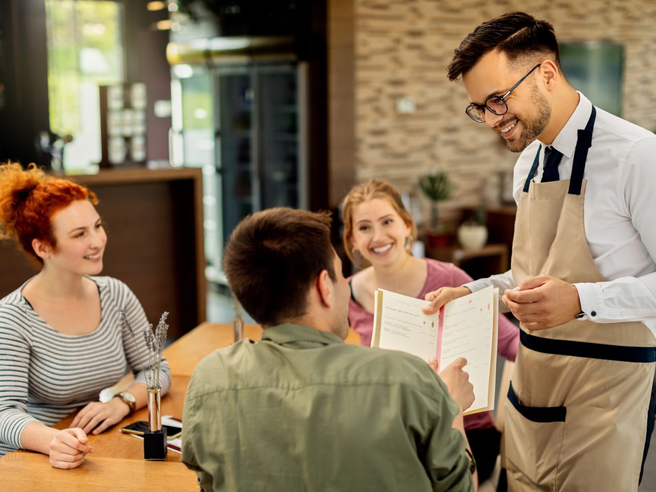 Happy waiter showing menu to a guest who came with friends in a cafe.