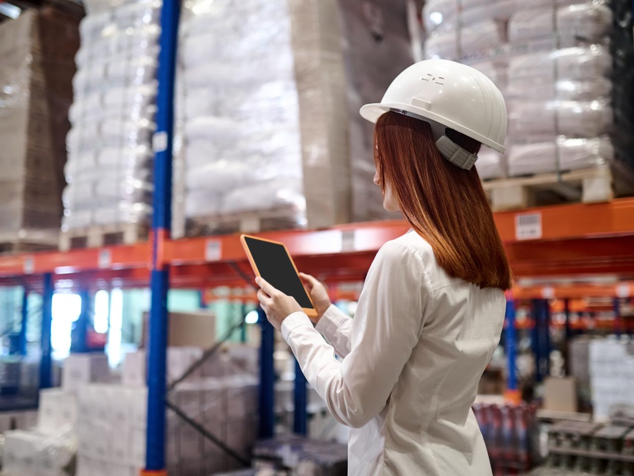 Management, warehouse. Profile of woman with long red hair in protective helmet looking at tablet screen walking along warehouse aisle