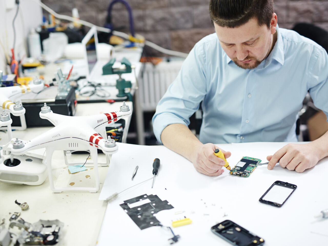 High angle shot of modern man repairing mobile device on table with different disassembled electronics, parts and drones in modern workshop