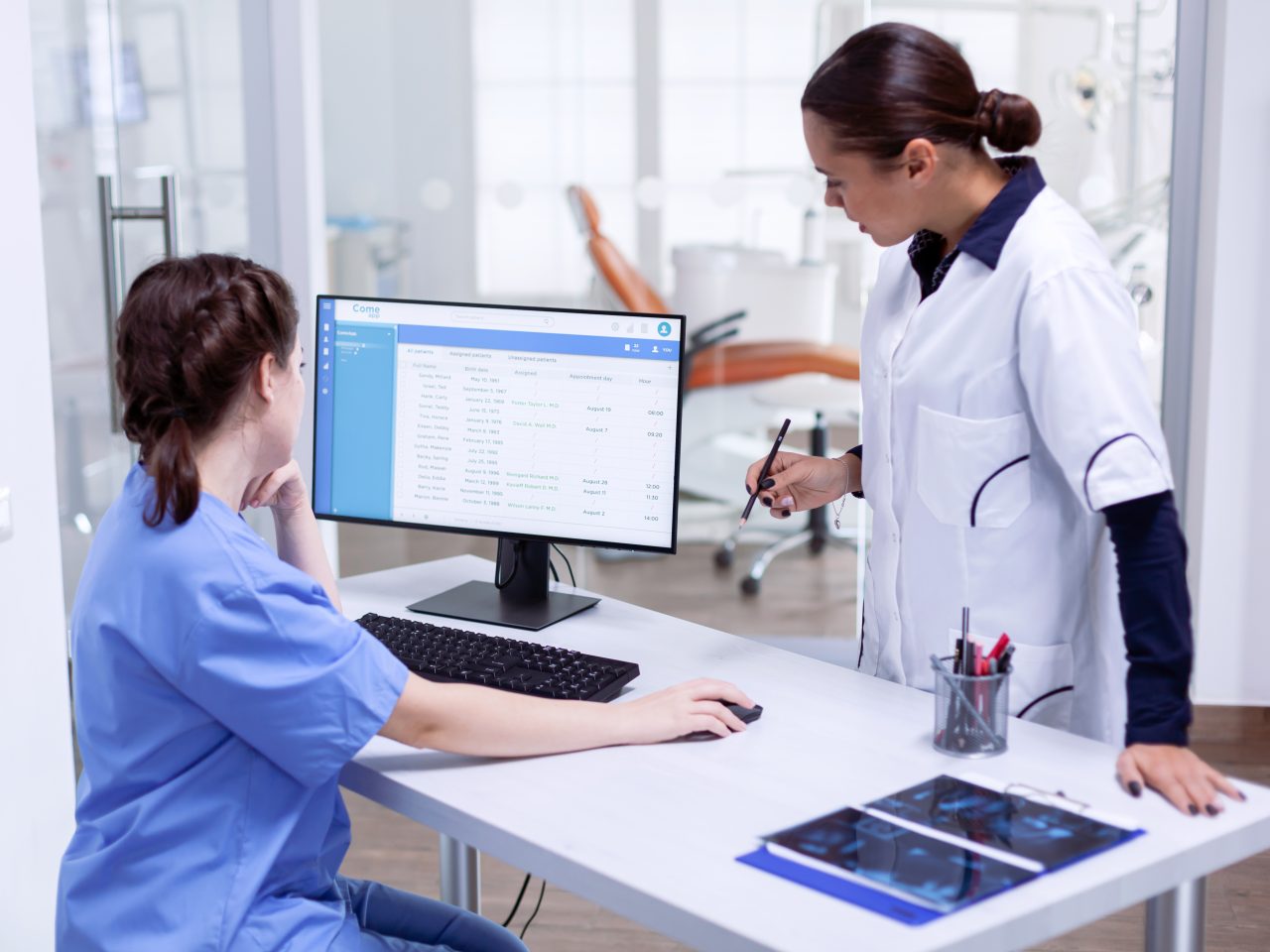 Stomatolog and nurse in tooth clinic checking patient appointment looking at computer monitor. Stomatology assistant and teeth doctor discussing in reception of dental office